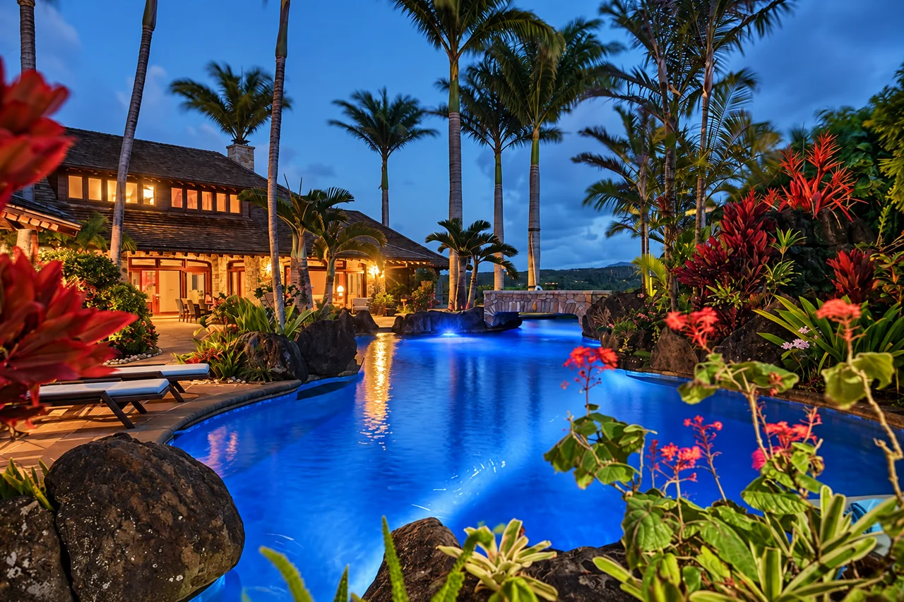 A blue-lit swimming pool surrounded by palms beside a Kauai villa at dusk