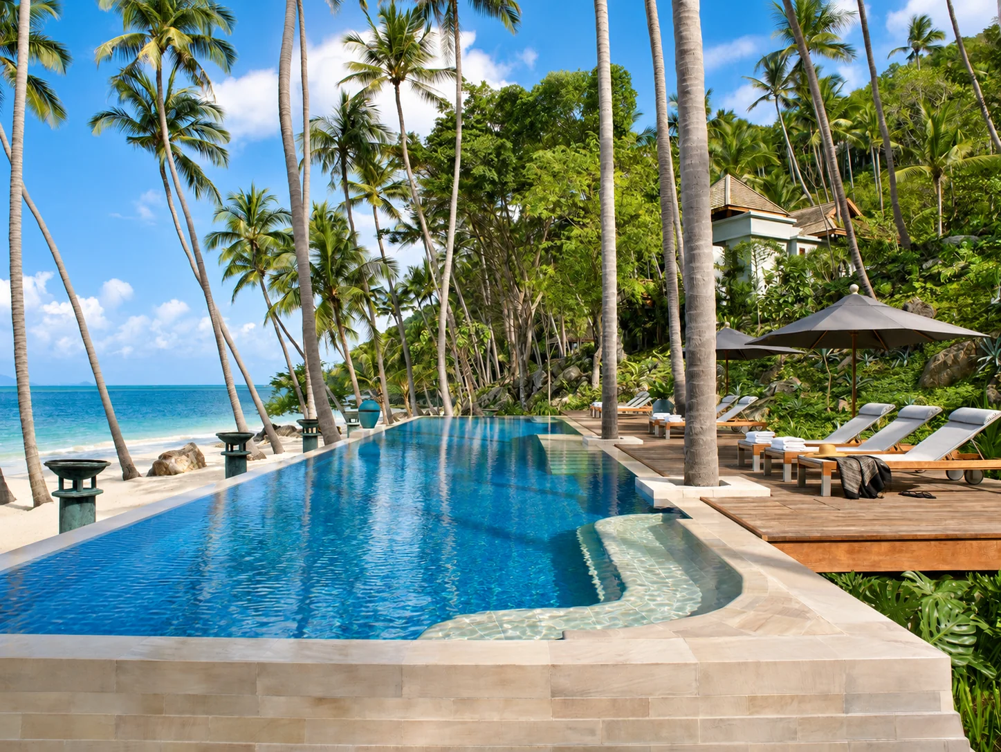 A beachfront infinity pool beside palms and a wooden lounge deck in Koh Samui