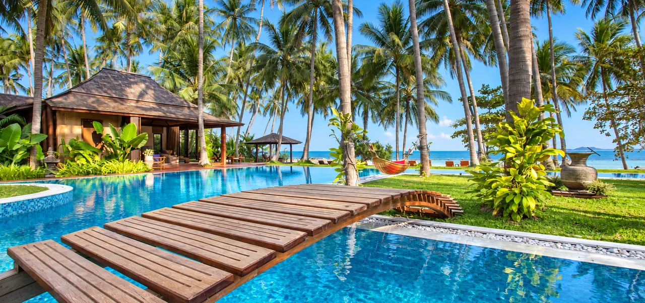 A wooden footbridge across a villa pool surrounded by coconut palms in Koh Samui